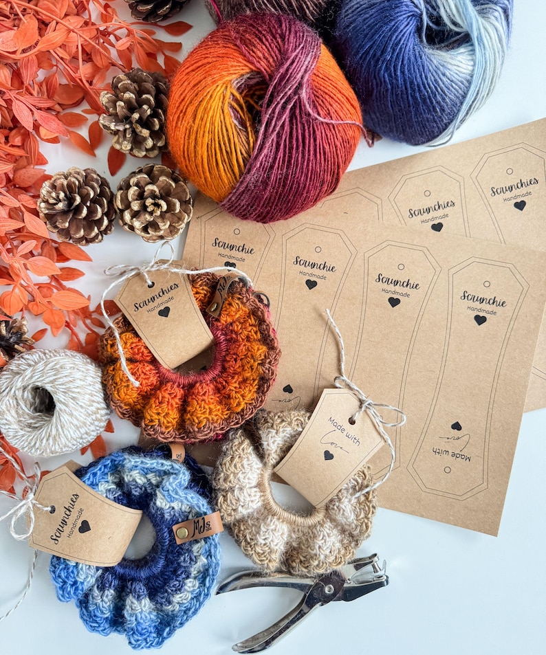 May include: An overhead shot features handmade scrunchies in shades of orange, blue, and beige, alongside skeins of yarn in complementary colors. The scrunchies have tags that read "Scrunchies Handmade" and "Made with Love". Pinecones and fall foliage add decorative elements.