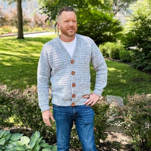 Op de afbeelding: Een lichtblauwe en witte gehaakte cardigan met houten knopen. De cardigan heeft een V-hals en lange mouwen. De persoon draagt een blauwe spijkerbroek en een wit t-shirt.