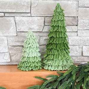 May include: Two green knitted Christmas trees of different sizes sit on a wooden surface. The larger tree is a darker green, while the smaller one is a lighter shade. A garland of green pine needles decorates the foreground.
