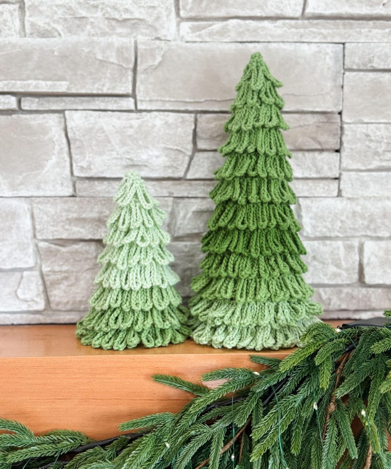 May include: Two green knitted Christmas trees of different sizes sit on a wooden surface. The larger tree is a darker green, while the smaller one is a lighter shade. A garland of green pine needles decorates the foreground.