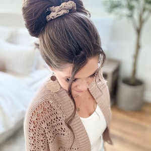 May include: A woman with brown hair styled in a high bun, secured with a beige crochet scrunchie. She wears a beige open-knit cardigan over a white top, complemented by a delicate gold necklace and round earrings. The overall aesthetic is soft and natural.