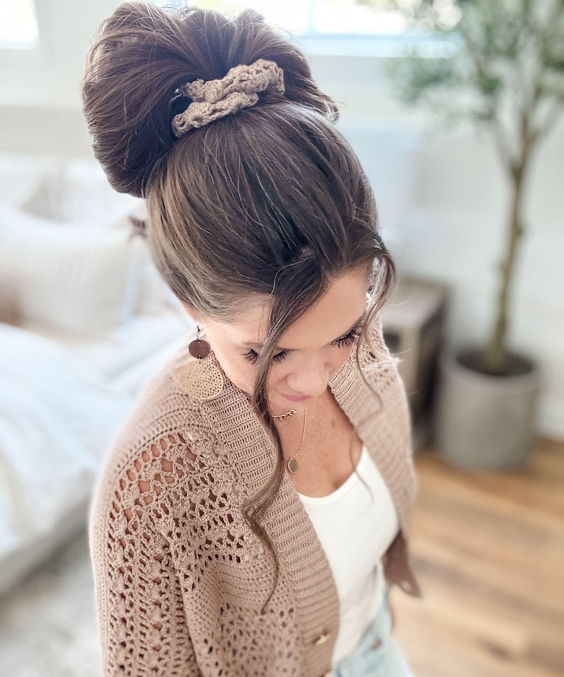 May include: A woman with brown hair styled in a high bun, secured with a beige crochet scrunchie. She wears a beige open-knit cardigan over a white top, complemented by a delicate gold necklace and round earrings. The overall aesthetic is soft and natural.
