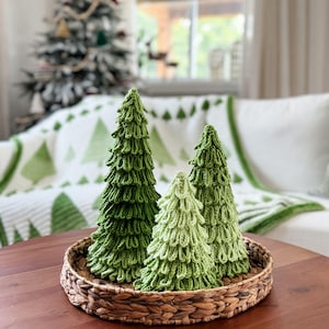May include: A wicker basket contains three knitted Christmas trees in shades of green. The trees feature a textured, layered design, mimicking evergreen foliage. The arrangement is on a dark wooden table, with a Christmas tree and a white sofa in the background.