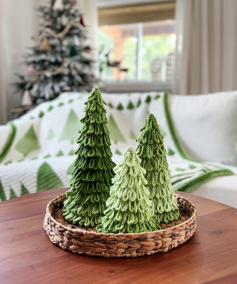 May include: A woven basket holds three knitted Christmas trees in varying shades of green. The trees have a textured, layered design, resembling evergreen foliage. The arrangement sits on a dark wooden table, with a Christmas tree and a white sofa in the background.