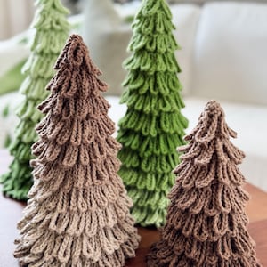 May include: Four handmade, knitted Christmas trees in varying shades of green and brown. The trees are arranged on a wooden surface, with a soft, blurred background. The trees have a textured, looped design, creating a unique, decorative look.