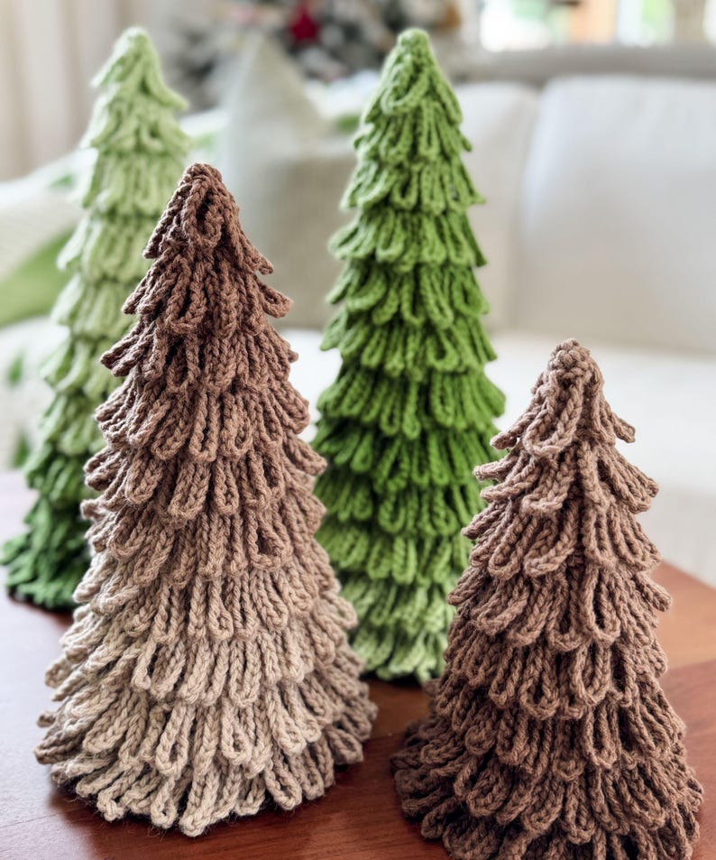 May include: Four handmade, knitted Christmas trees in varying shades of green and brown. The trees are arranged on a wooden surface, with a soft, blurred background. The trees have a textured, looped design, creating a unique, decorative look.