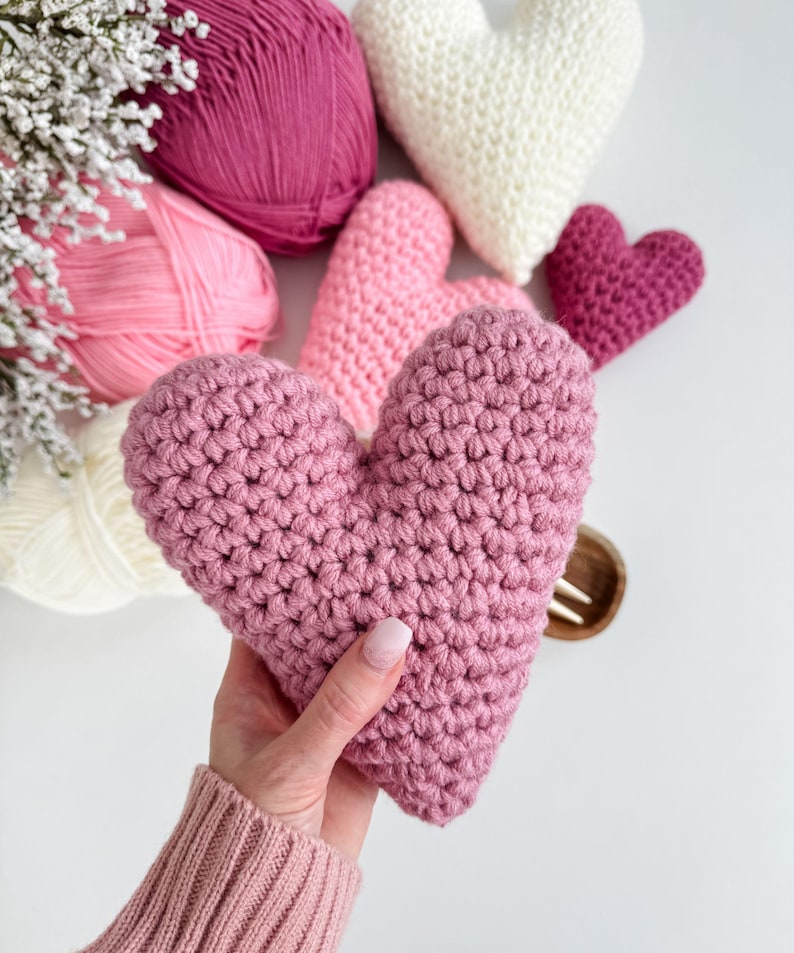 Pu&ograve; includere: Una collezione di cuori all'uncinetto rosa e bianchi, alcuni sono gomitoli di lana e altri hanno la forma di un cuore. I cuori sono su una superficie bianca.