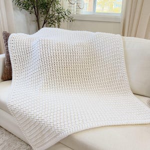 Può includere: Una coperta bianca all'uncinetto e strutturata drappeggiata su un divano bianco. La coperta ha un motivo intrecciato e un bordo spesso. Un cuscino marrone è visibile sul lato sinistro del divano. Lo sfondo include una finestra, tende e una pianta decorativa.