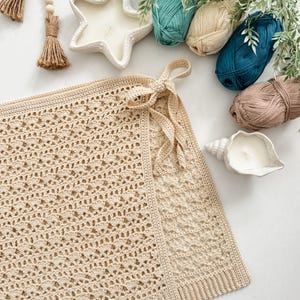 May include: A beige crochet blanket with a delicate openwork pattern, tied with a bow. The image also features yarn in various shades of blue and brown, a starfish-shaped candle, and a seashell candle holder.