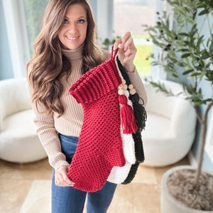 May include: A person holding three handmade Christmas stockings. The stockings are red, white, and black, with decorative tassels and wooden beads. The red stocking is in the front, with a folded cuff. The stockings are made of a knitted material.
