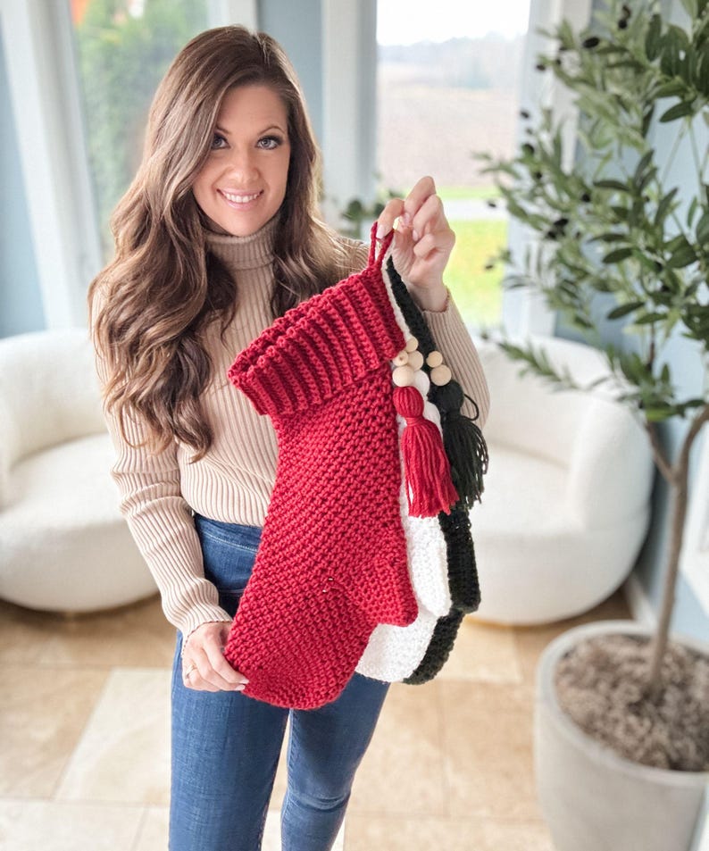 May include: A person holding three handmade Christmas stockings. The stockings are red, white, and black, with decorative tassels and wooden beads. The red stocking is in the front, with a folded cuff. The stockings are made of a knitted material.