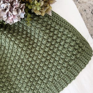 May include: Close-up of a sage green knitted blanket with a textured, raised pattern. The blanket is partially visible, with a portion of the edge and the textured surface in focus. Dried flowers in shades of pink, green, and brown are in the background.