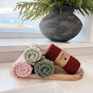 May include: Rolled crochet dishcloths in shades of pink, green, and burgundy, displayed on a wooden tray. A vase with greenery sits in the background. The burgundy dishcloth has a paper label that reads "Dishcloth Handmade".