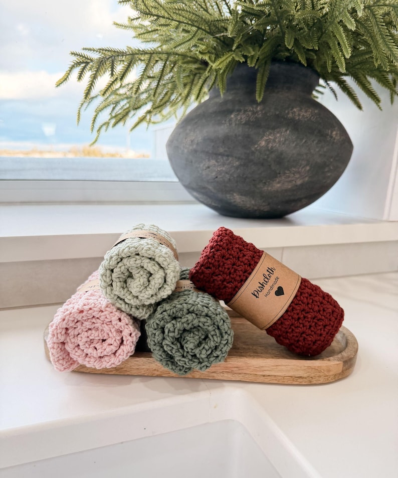 May include: Rolled crochet dishcloths in shades of pink, green, and burgundy, displayed on a wooden tray. A vase with greenery sits in the background. The burgundy dishcloth has a paper label that reads "Dishcloth Handmade".