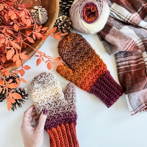 May include: Handmade crochet mittens in shades of brown, orange, and burgundy. The mittens feature a textured stitch pattern and ribbed cuffs. A ball of yarn and a plaid scarf are also in the image, suggesting a cozy, autumn theme.
