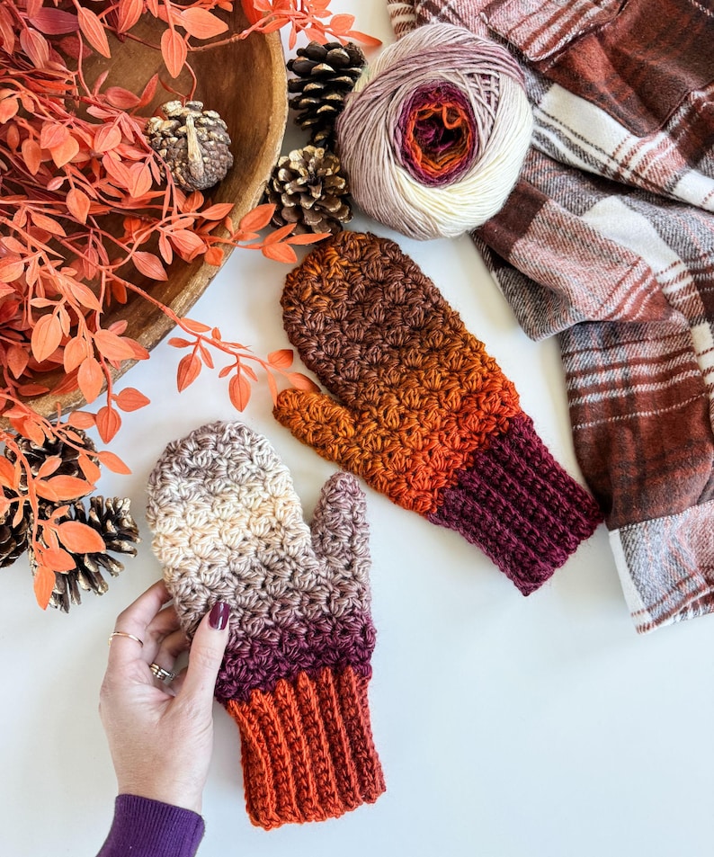 Peut inclure: Mitaines crochet&eacute;es &agrave; la main, aux tons bruns, orange et bordeaux. Les mitaines pr&eacute;sentent un motif de point textur&eacute; et des poignets c&ocirc;tel&eacute;s. Une pelote de laine et une &eacute;charpe &agrave; carreaux sont &eacute;galement dans l'image, sugg&eacute;rant un th&egrave;me automnal et chaleureux.