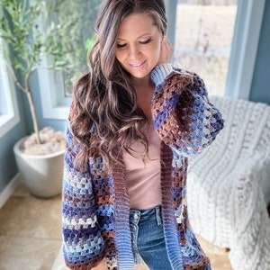 K&ouml;nnte beinhalten: Ein langer, geh&auml;kelter Cardigan mit braunen, blauen und wei&szlig;en Streifen. Der Cardigan hat eine lockere Passform und einen entspannten Stil.