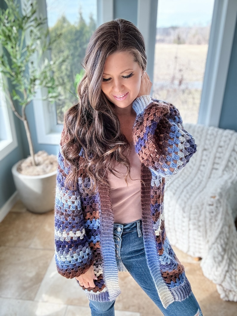 K&ouml;nnte beinhalten: Ein langer, geh&auml;kelter Cardigan mit braunen, blauen und wei&szlig;en Streifen. Der Cardigan hat eine lockere Passform und einen entspannten Stil.
