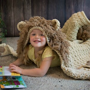 May include: A young child wearing a crocheted lion costume with a brown and beige fur hood and a matching blanket. The child is lying on a light brown surface and reading a book.