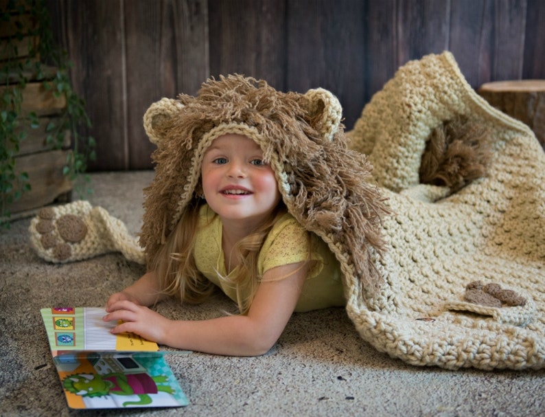 May include: A young child wearing a crocheted lion costume with a brown and beige fur hood and a matching blanket. The child is lying on a light brown surface and reading a book.