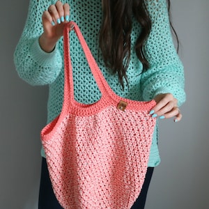 May include: A crocheted pink and white tote bag with a brown leather tag. The bag is being held by a person wearing a turquoise crochet sweater.