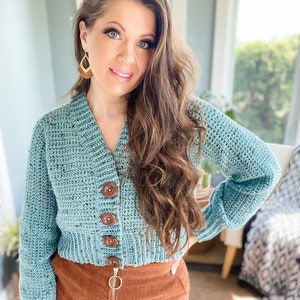 Op de afbeelding: Een tealblauwe gehaakte cardigan met bruine knopen. De cardigan heeft een v-hals en lange mouwen. Het model draagt een bruine corduroy rok.