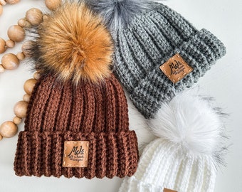 May include: Three crocheted beanies with faux fur pom poms. The beanies are brown, gray, and white. Each beanie has a leather patch with the text "Mel's Off the Hook Designs".