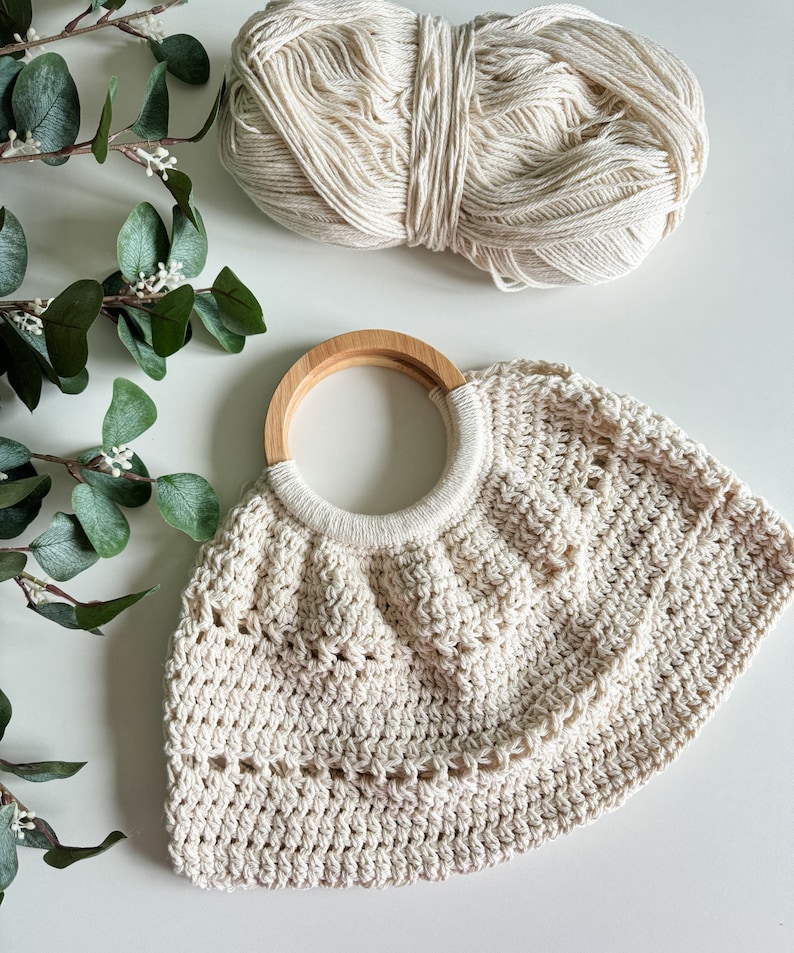 May include: A crocheted cream-colored tote bag with a wooden handle. The bag has a scalloped edge and a delicate openwork pattern.