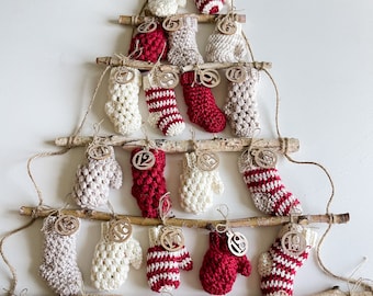 May include: A Christmas advent calendar made of crocheted mittens in red, white, and cream colors. Each mitten has a numbered wooden tag attached to it. The mittens are hung on a wooden branch structure.