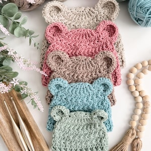 Puede incluir: Una pila de cinco gorros tejidos a mano al crochet, cada uno con dos pequeñas orejas de oso redondeadas en la parte superior. Los gorros están dispuestos en una fila vertical, mostrando colores beige, rosa empolvado, marrón, azul cerceta y verde salvia.