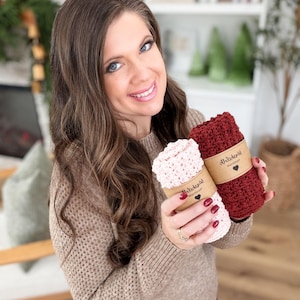 May include: A woman holds two rolled-up crochet washcloths. One is light pink, the other is burgundy. Each is wrapped with a brown paper band featuring a heart and text. The woman is wearing a tan sweater.