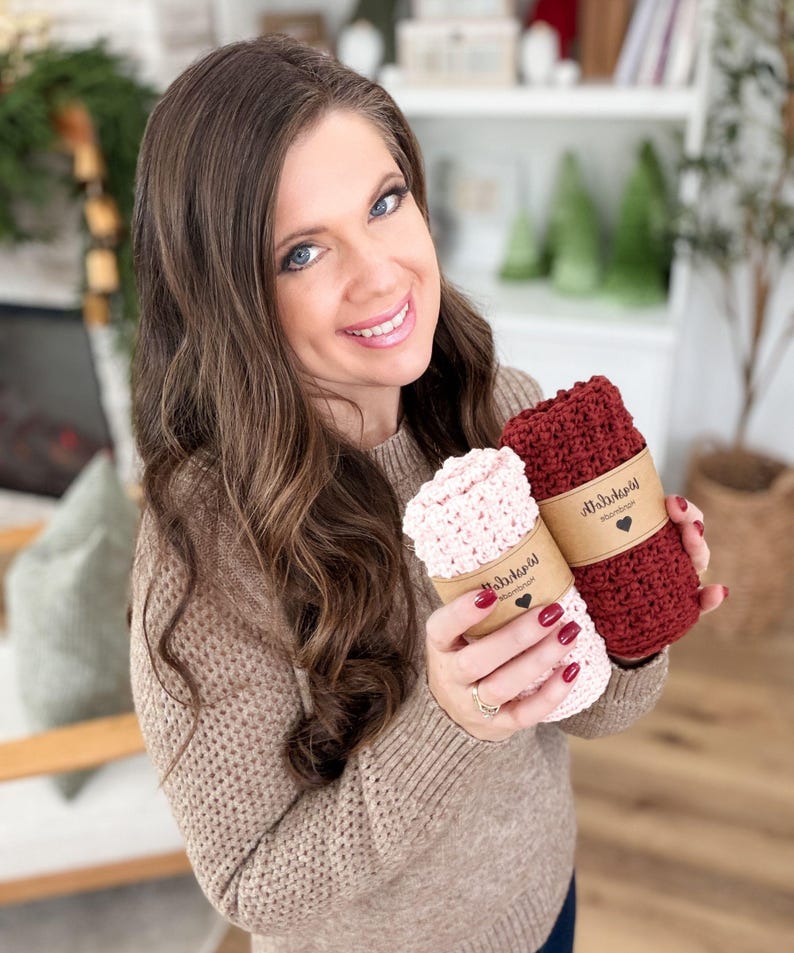 May include: A woman holds two rolled-up crochet washcloths. One is light pink, the other is burgundy. Each is wrapped with a brown paper band featuring a heart and text. The woman is wearing a tan sweater.