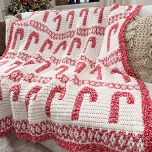 K&ouml;nnte beinhalten: Eine wei&szlig;-rote H&auml;keldecke mit einem sich wiederholenden Zuckerstangenmuster. Die Decke mit rotem Rand ist &uuml;ber ein Sofa drapiert. Diese Weihnachtsdecke ist ein gem&uuml;tliches Wohnaccessoire f&uuml;r die festliche Jahreszeit.