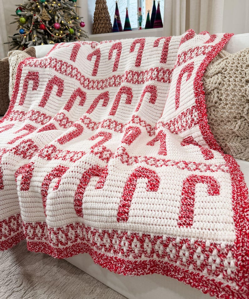 K&ouml;nnte beinhalten: Eine wei&szlig;-rote H&auml;keldecke mit einem sich wiederholenden Zuckerstangenmuster. Die Decke mit rotem Rand ist &uuml;ber ein Sofa drapiert. Diese Weihnachtsdecke ist ein gem&uuml;tliches Wohnaccessoire f&uuml;r die festliche Jahreszeit.
