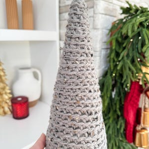 May include: A gray knitted cone-shaped Christmas tree decoration. The tree is made of a chunky yarn with a speckled texture.