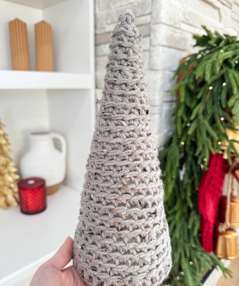 May include: A gray knitted cone-shaped Christmas tree decoration. The tree is made of a chunky yarn with a speckled texture.