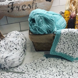 May include: Two balls of yarn, one teal and one white with black flecks, are in a wire basket with a burlap liner. A teal crocheted dishcloth is on a white and black crocheted dishcloth.