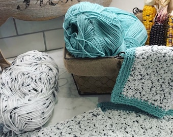 May include: Two balls of yarn, one teal and one white with black flecks, are in a wire basket with a burlap liner. A teal crocheted dishcloth is on a white and black crocheted dishcloth.