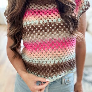 May include: A colorful crochet top with a gradient design. The top features a mix of pink, brown, and beige tones, creating a striped pattern. The short-sleeved top has a round neckline and a scalloped hemline, perfect for a summer look.
