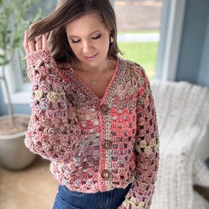 May include: A crocheted cardigan with a multi-colored pattern in shades of pink, peach, and cream. The cardigan has a V-neck and button closure. The sleeves are long and have a loose fit.