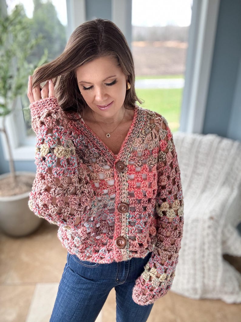 May include: A crocheted cardigan with a multi-colored pattern in shades of pink, peach, and cream. The cardigan has a V-neck and button closure. The sleeves are long and have a loose fit.