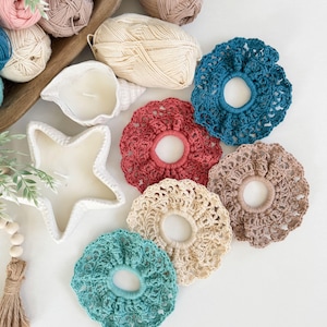 May include: A collection of handmade crochet hair scrunchies in various colors, including teal, red, and beige. The scrunchies are arranged on a white surface with yarn balls and decorative candles in the background. The scrunchies are a mix of solid and patterned designs.