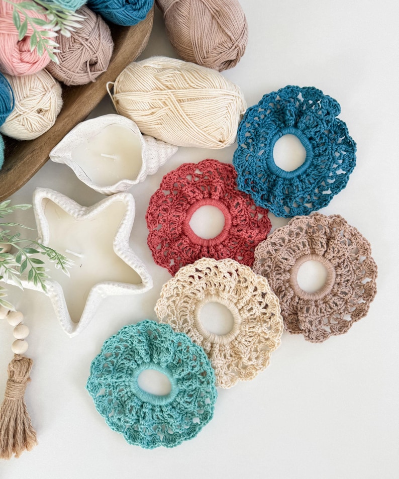 May include: A collection of handmade crochet hair scrunchies in various colors, including teal, red, and beige. The scrunchies are arranged on a white surface with yarn balls and decorative candles in the background. The scrunchies are a mix of solid and patterned designs.