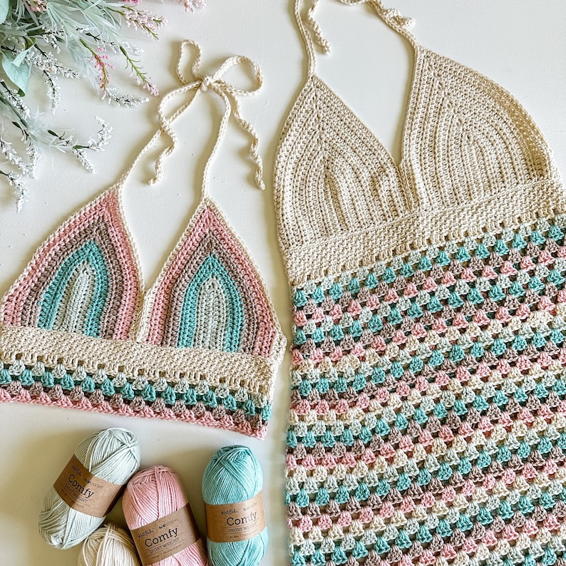 May include: A crochet bikini top and matching dress in a light brown, pink, and teal color scheme. The top is a halter style with a triangle shape and the dress is a halter style with a square neckline. The dress has a geometric pattern.