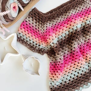 May include: A handmade crochet top in a granny square pattern, featuring a palette of brown, pink, and light blue hues. The top is displayed on a white surface, alongside a wooden bowl of yarn and decorative candles.