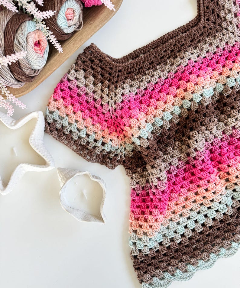 May include: A handmade crochet top in a granny square pattern, featuring a palette of brown, pink, and light blue hues. The top is displayed on a white surface, alongside a wooden bowl of yarn and decorative candles.