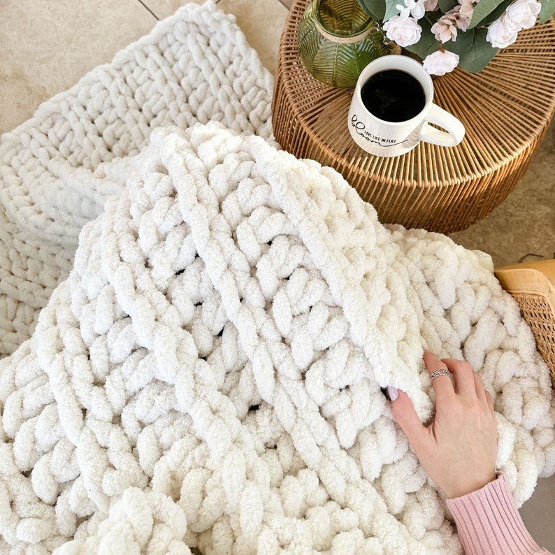 Peut inclure: Une couverture en tricot blanc &eacute;pais avec une texture douce et moelleuse. La couverture est drap&eacute;e sur une chaise et une table avec une tasse de caf&eacute; dessus.