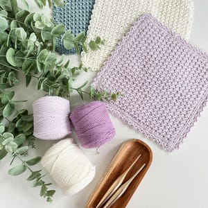 May include: Three skeins of yarn in shades of lavender, white, and light blue, along with two crochet hooks in a wooden bowl. The yarn is arranged on a white surface with a sprig of eucalyptus.