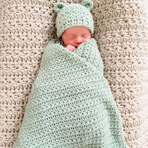 Puede incluir: Un reci&eacute;n nacido est&aacute; envuelto en una manta de ganchillo de color verde salvia claro con un patr&oacute;n de punto de estrella. El reci&eacute;n nacido lleva un gorro de ganchillo a juego de color verde salvia claro con dos orejas de oso.