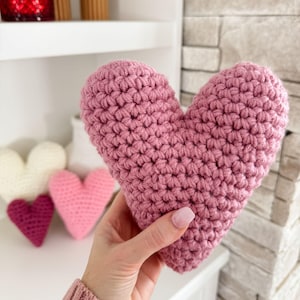 Pu&ograve; includere: Un cuscino a forma di cuore in crochet rosa. Il cuscino &egrave; tenuto in una mano.